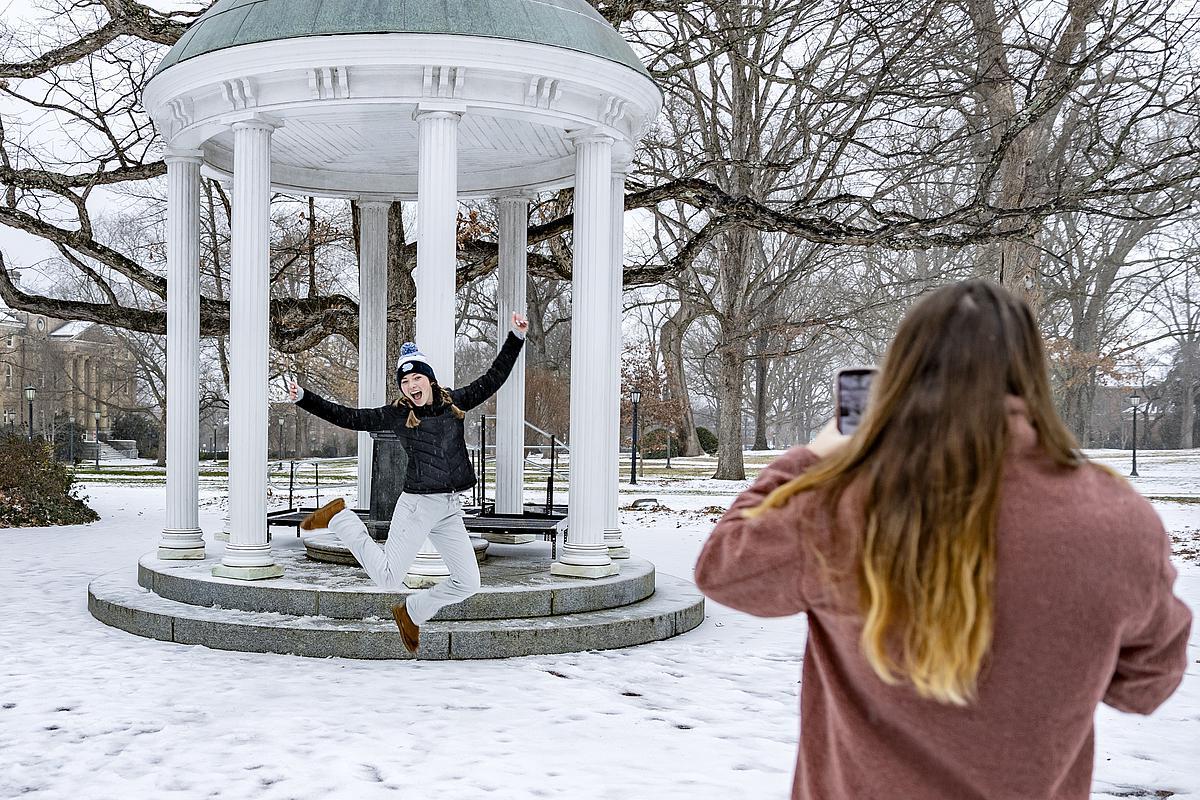 A student standing in the foreground taking a photo of her friend leaping in front the Old Well during the winter season.